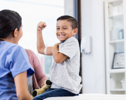 A young boy smiles and shows his biceps to a doctor. He has an adhesive bandage on his arm.