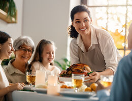 A smiling woman places a roasted turkey in the middle of a family dinner table.